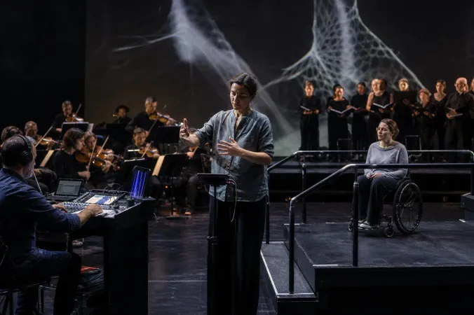 A nonspeaking performer on an opera stage using a small motion-control device as musicians and singers perform nearby.