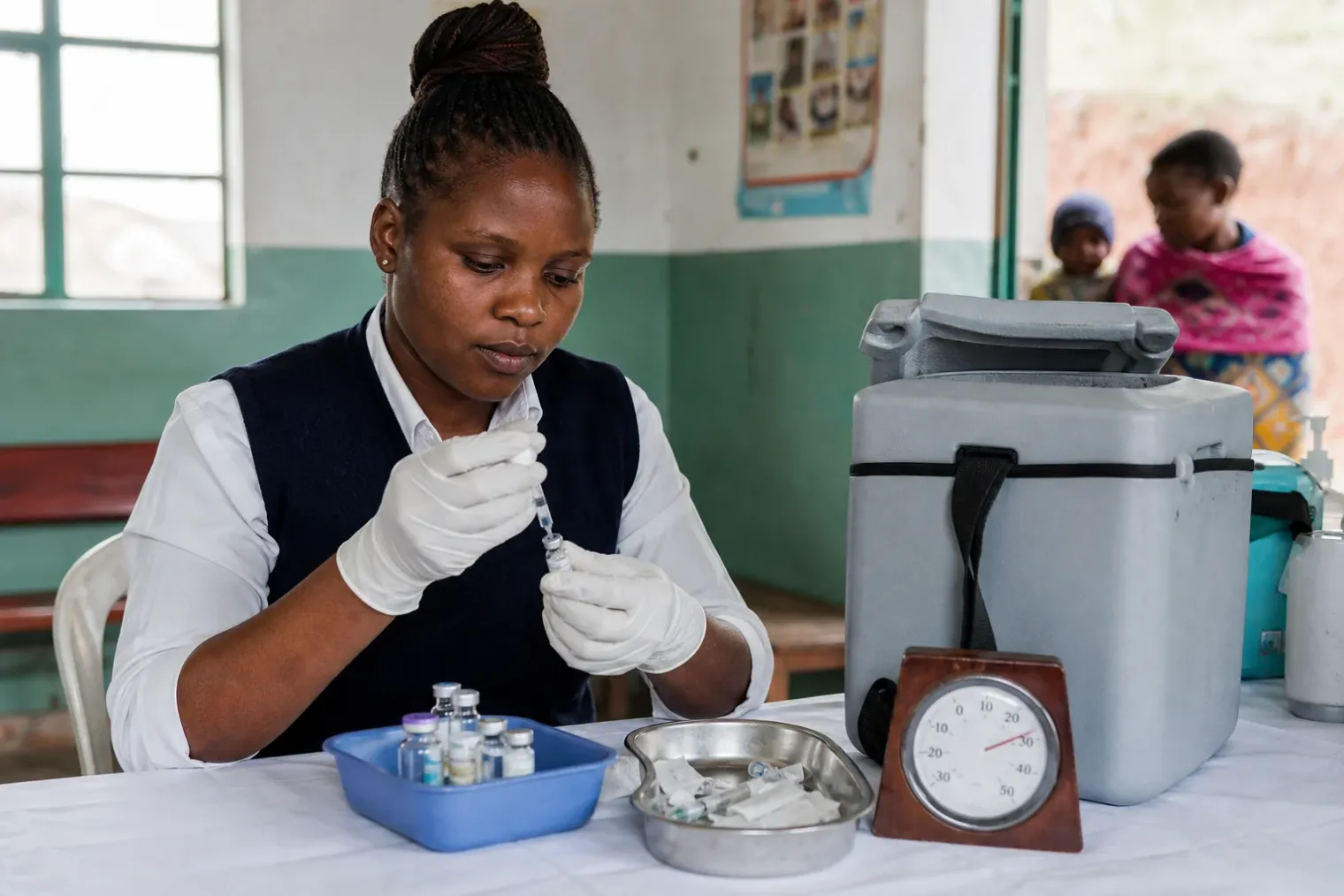 A healthcare worker preparing a vaccine vial with sterile water beside a thermometer in a rural clinic without refrigeration equipment.