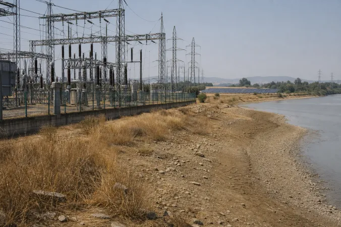 A European electricity transmission substation beside a low river during a summer heatwave, with solar panels in the distance.