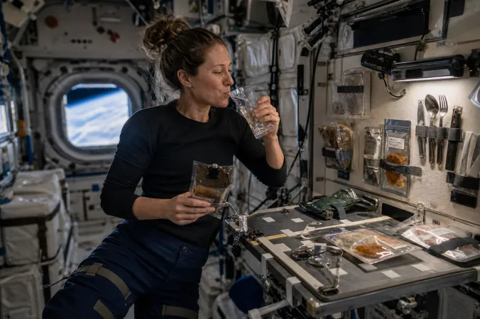 An astronaut floating at a meal station inside a spacecraft, drinking from a sealed pouch and holding a secured food container.
